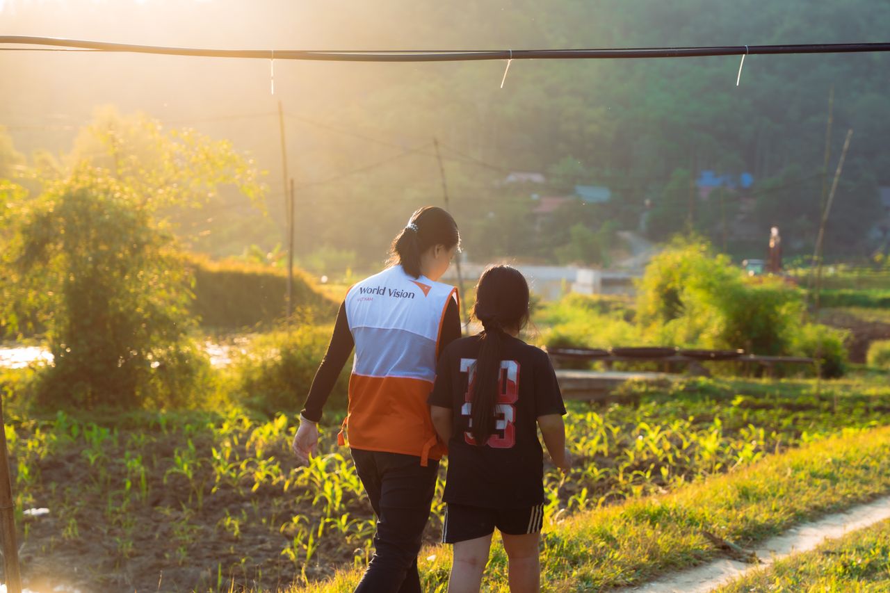 hero-img-02 Volunteer in World Vision vest walking with child in scenic rural landscape at sunset.