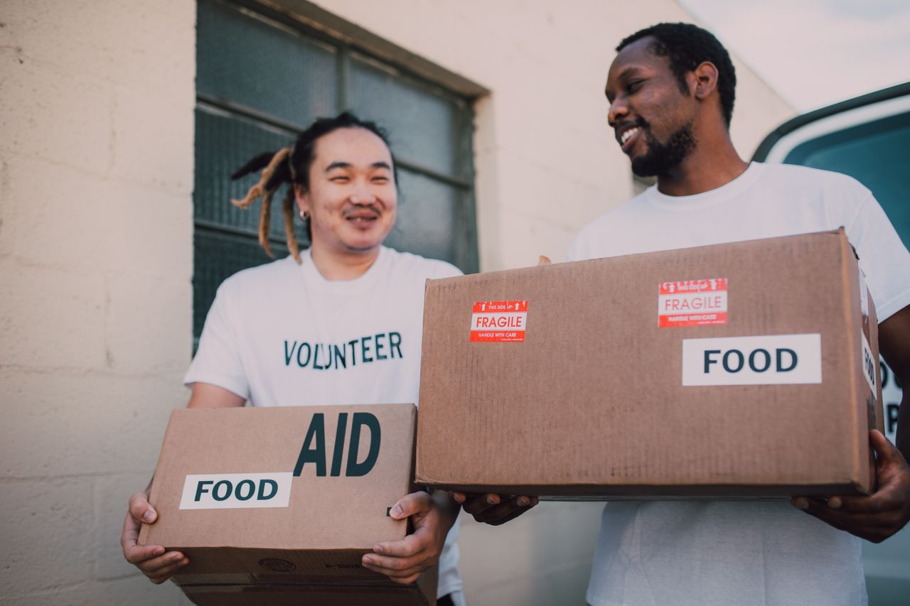hero-img-01 Two volunteers carrying aid boxes labeled food, showing community support and charity efforts.